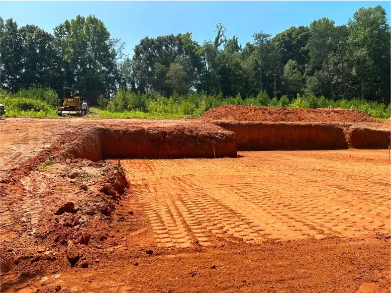 Construction site with red dirt, leveled ground, and a small excavator. Trees in the background.