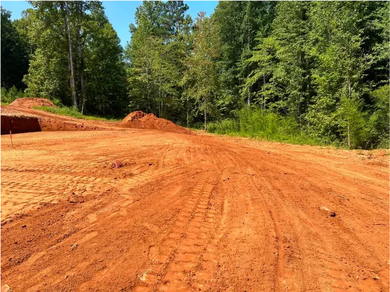 Dirt clearing with tire tracks, piles of dirt, and a backdrop of green trees under a blue sky.