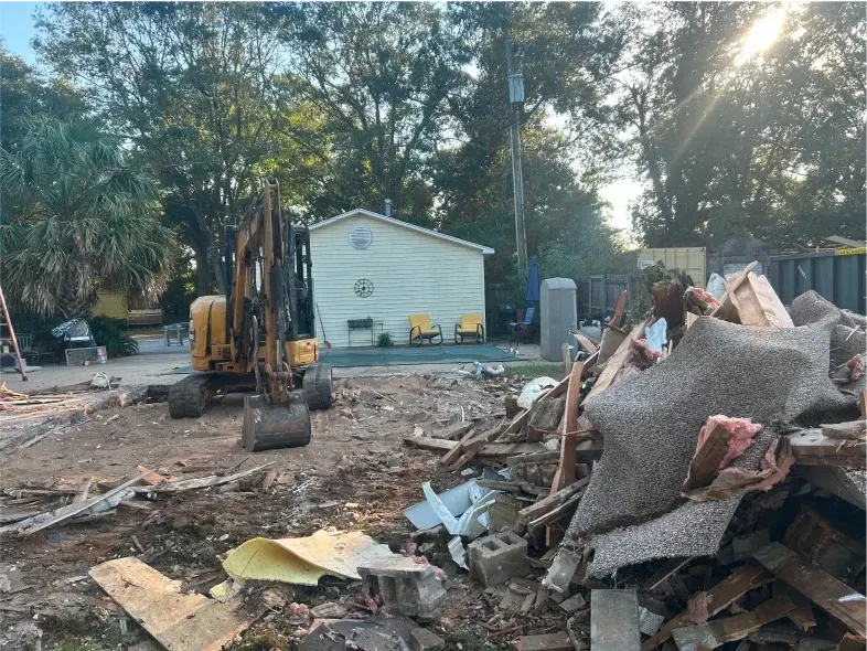 Demolition site with excavator, debris pile, and small building in the background.