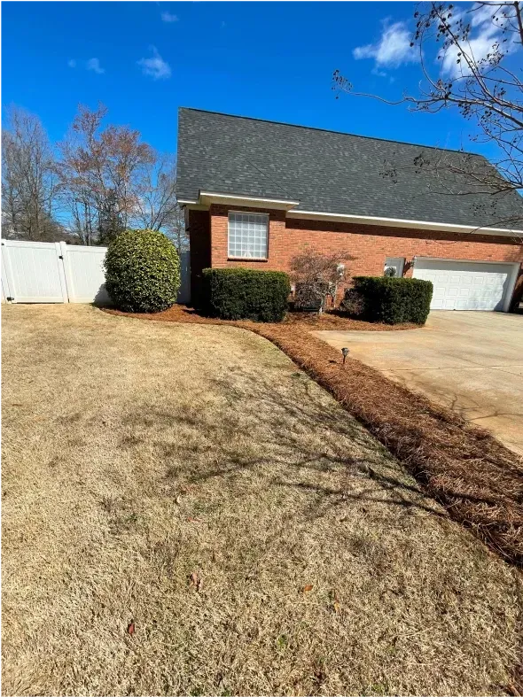 Exterior of a brick house with a white fence, green bushes, and a lawn covered in brown grass.