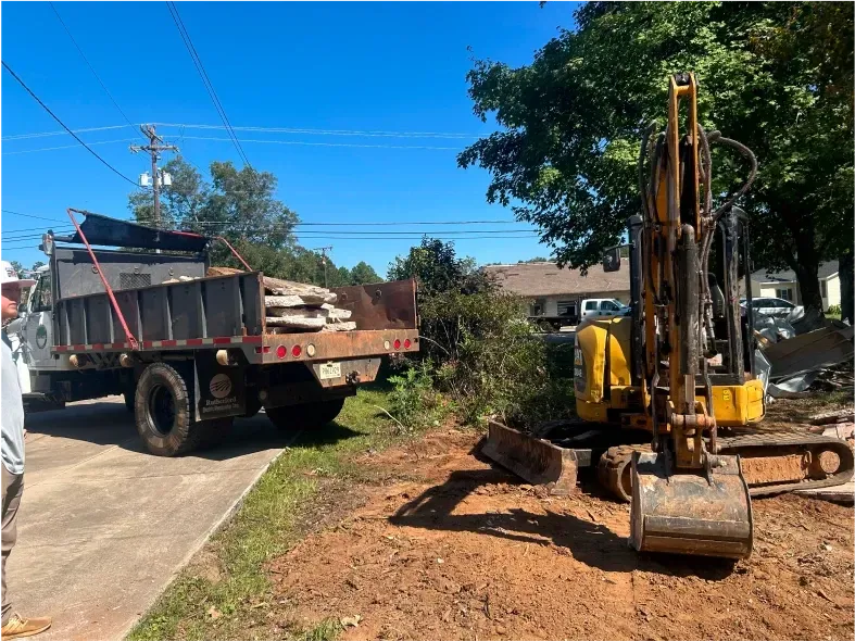 A yellow excavator next to a dump truck loaded with concrete slabs on a construction site.
