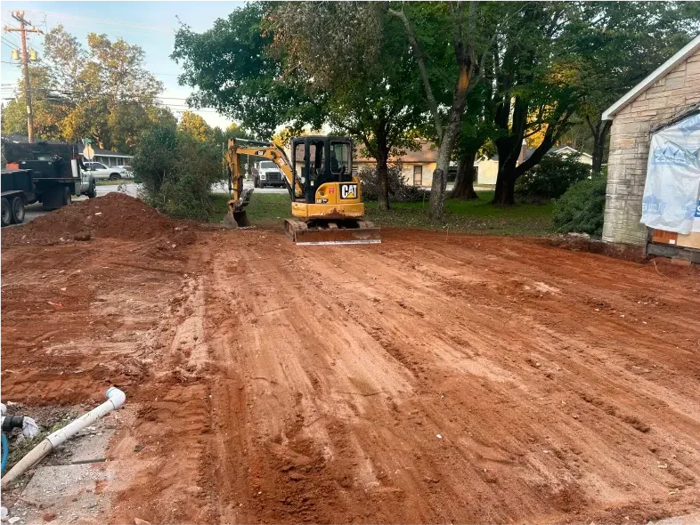 Excavator compacting soil at a construction site next to a building, with a truck and trees in the background.