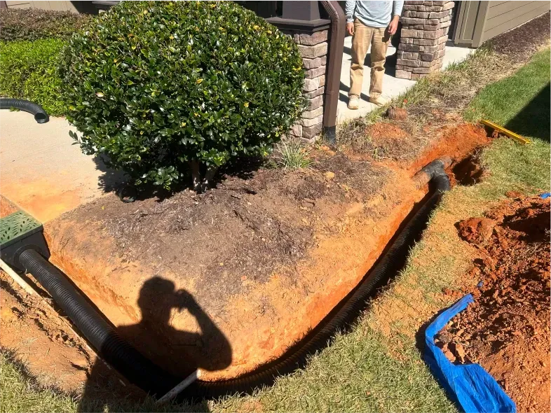 Trench with black pipe installed near a bush, person standing nearby. Ground is reddish-brown.