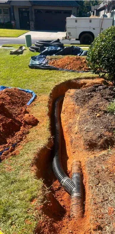 Drainage pipe installed in a trench on a residential lawn.  Red soil, green grass, and a white utility truck are in the background.