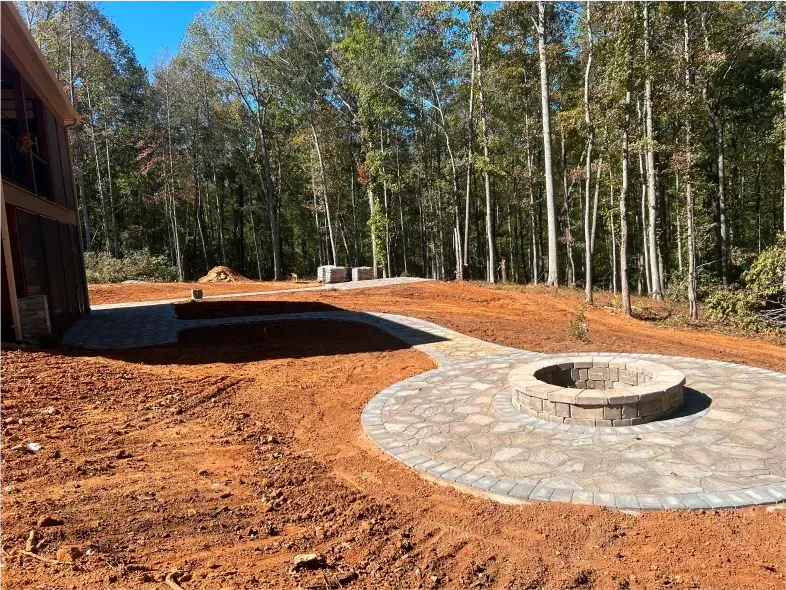 A circular stone fire pit with a pathway on a dirt lot, surrounded by trees.