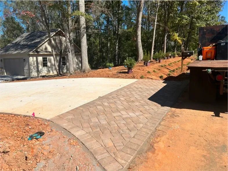 Driveway with brick-like paving curving into a concrete driveway. A garage sits on the left, trees in the background.