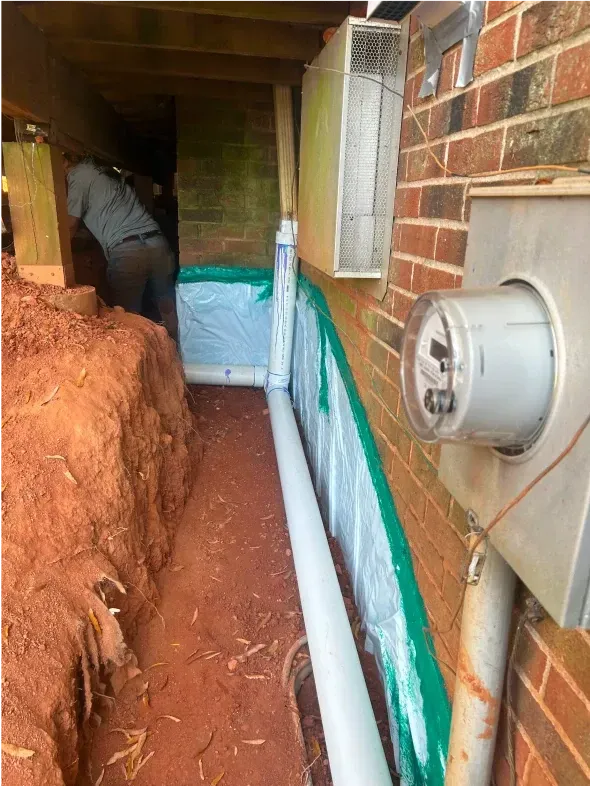Construction site with a trench next to a brick wall. A worker is in the background. Drain pipe visible.