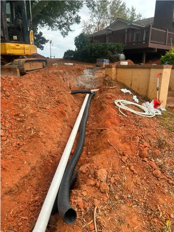 Trench in red dirt with white and black pipes, an excavator, and a house in the background.