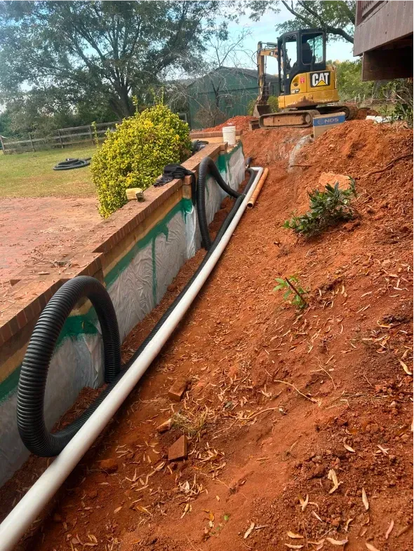 Construction site with drainage pipes and a small excavator. Red dirt surrounds a retaining wall.