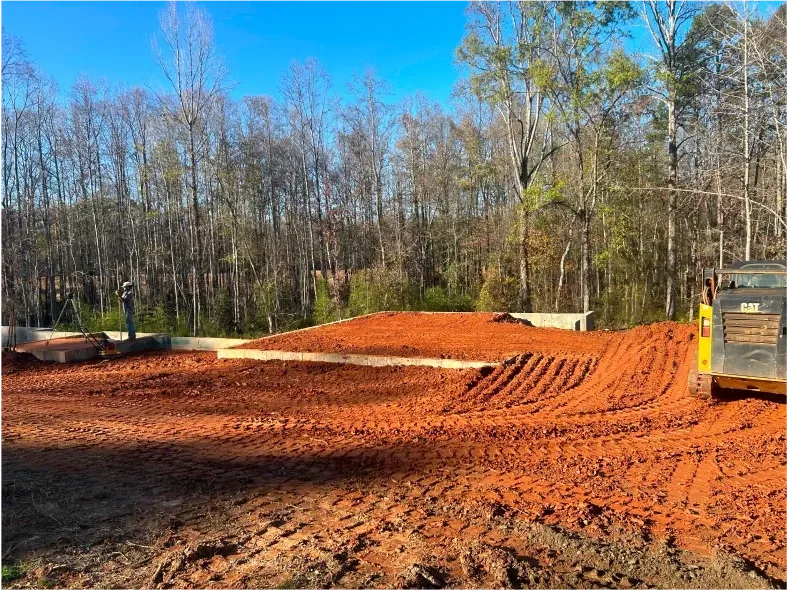 Construction site with red dirt graded by a bulldozer, in front of a forest.