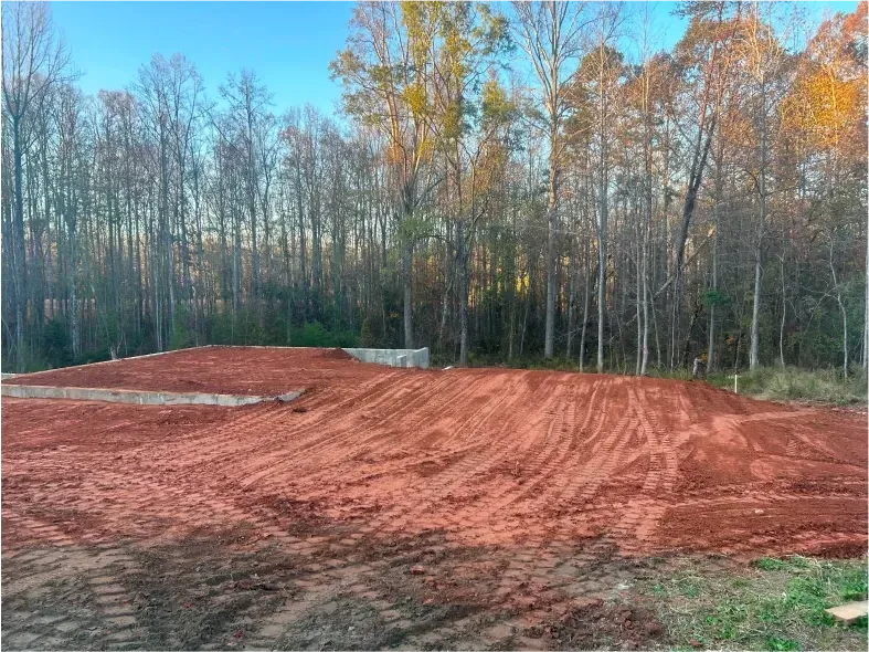 A red dirt lot leveled for construction, with trees in the background under a blue sky.