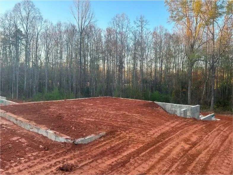 Construction site with red dirt, concrete foundation, and bare trees in the background.