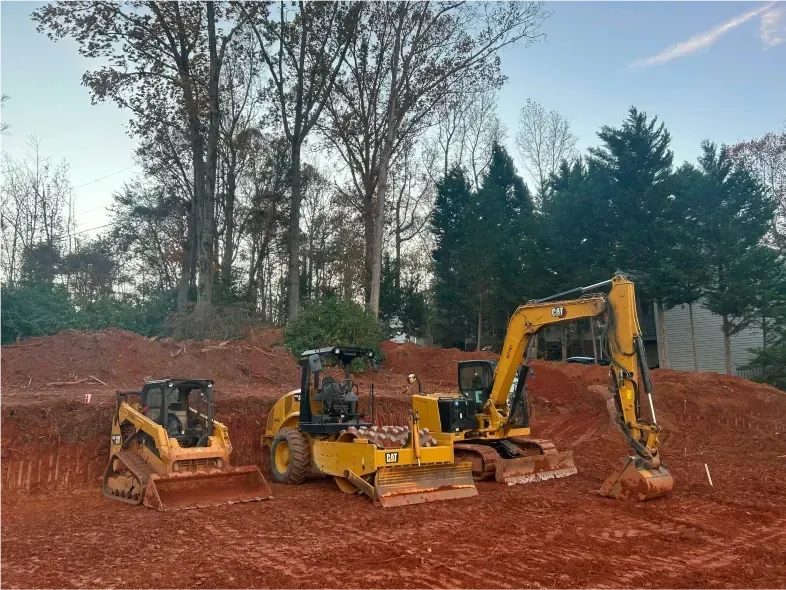 Construction site with three Caterpillar machines on red dirt; trees and sky in the background.