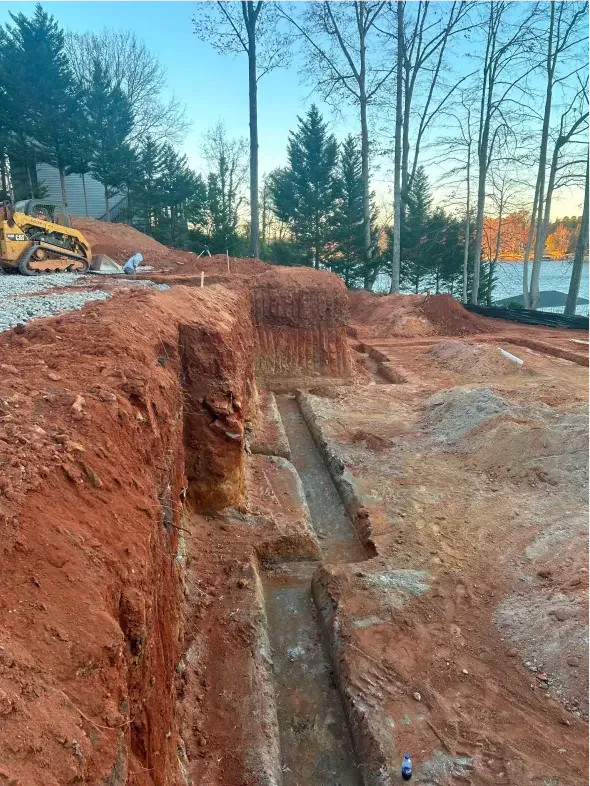 Construction site with excavated foundation trenches in red soil, with trees and water in the background.