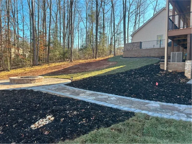 Backyard with stone path, fire pit, and house with landscaping on a hill. Black mulch, grass, and trees.