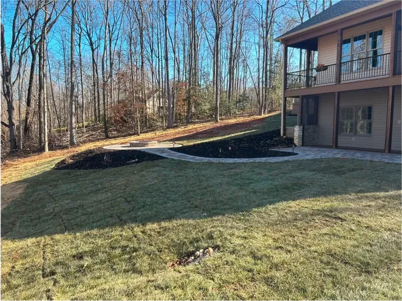 Backyard with freshly mown lawn, stone patio, and two-story house with balcony overlooking a wooded area.