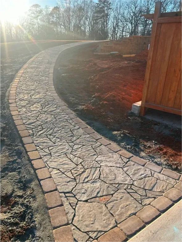 Stone pathway winding upward, bordered by brick and earth, beside a wooden structure and sunny hillside.