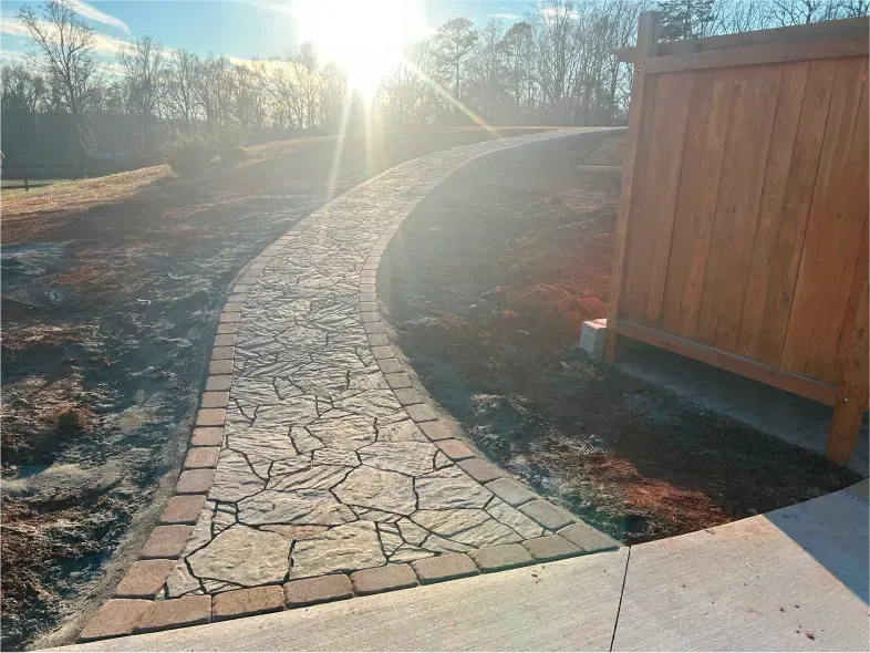 Stone walkway curving past a wooden fence and into sunlight.