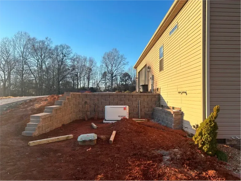 Residential building exterior with a retaining wall, steps, and generator on a mound of mulch.