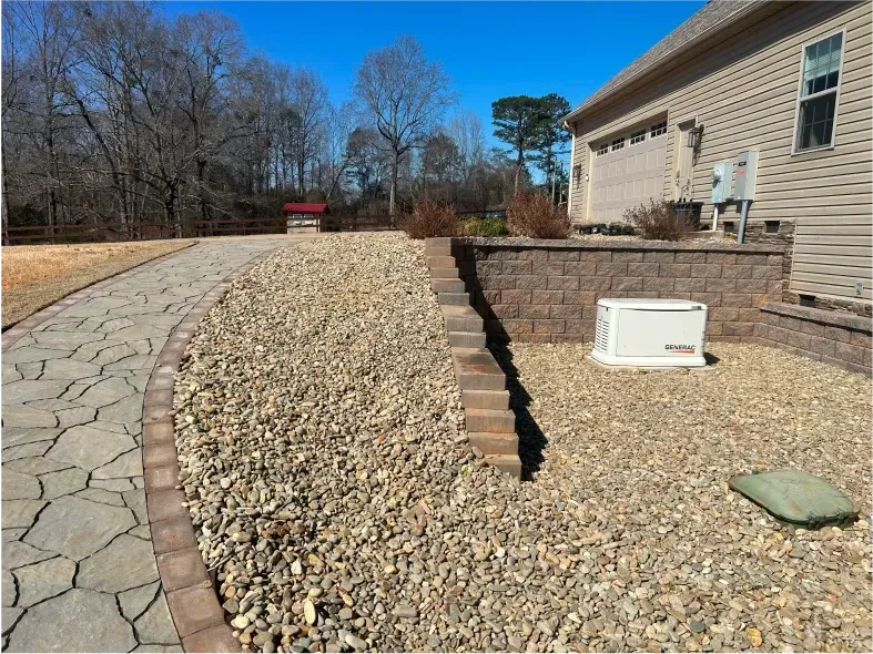 Stone pathway curves alongside a gravel bed with a retaining wall, a generator, and a house.