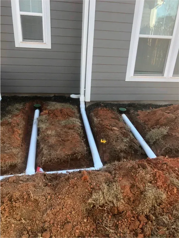 Three white pipes in trenches near a house, connected to a downspout.