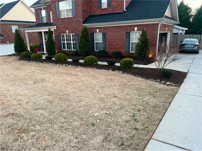 Two-story brick house with a brown mulch bed, green shrubs, and a driveway with a parked car.