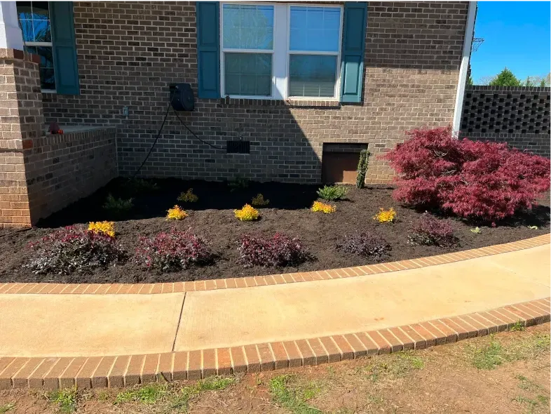 Brick building with flower bed containing plants and a walkway bordered by bricks.