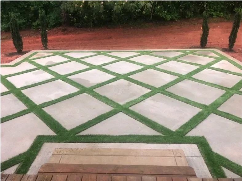Concrete patio with diamond-patterned artificial turf inlays; cypress trees and red ground in the background.