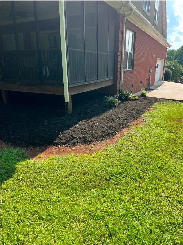 Freshly mulched garden bed along a house with screened porch and brick exterior, next to green lawn.