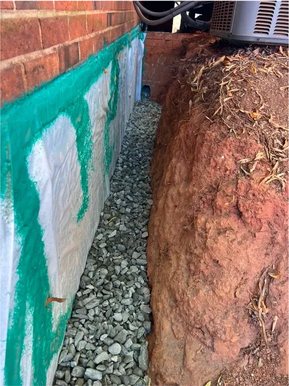 Foundation drainage system: gravel-filled trench between brick wall (green coating) and exposed soil.