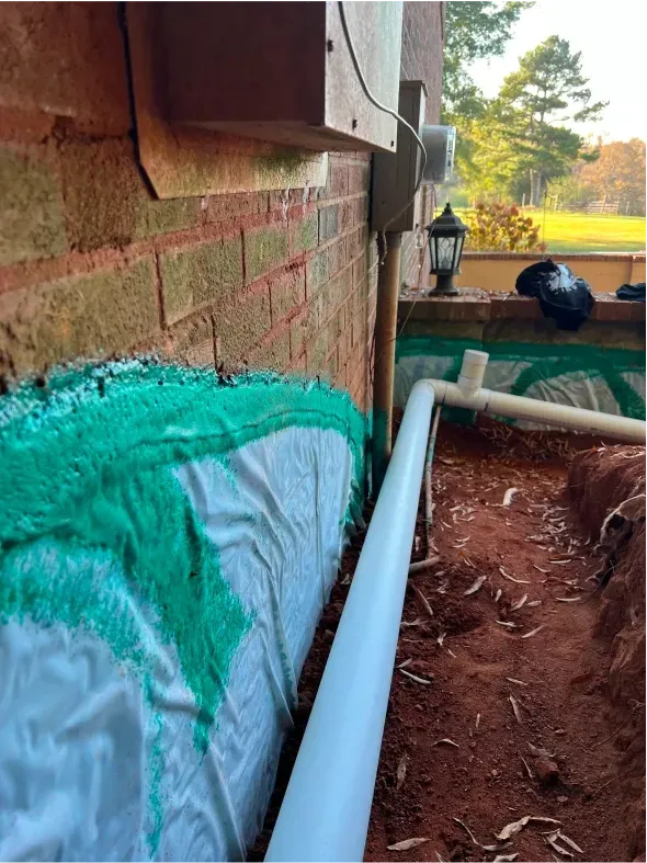 Exterior view of a brick wall, green mesh, white fabric, and a PVC pipe in a dirt trench, likely for drainage.