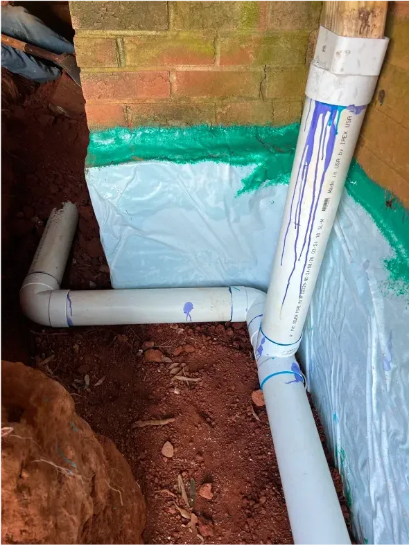 PVC pipes exiting a brick foundation wall, connected with elbow joints. Blue paint on pipes.