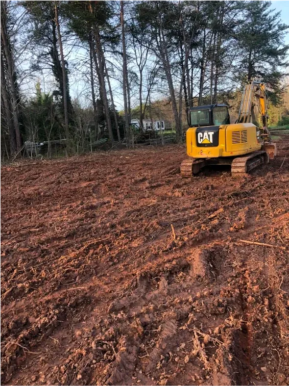 Yellow CAT excavator on a freshly tilled, red-brown earth clearing site, trees in background.