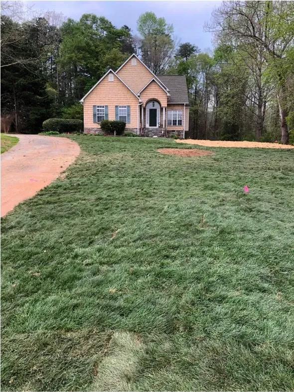 House with a lawn, driveway on the left, trees in background, cloudy sky.