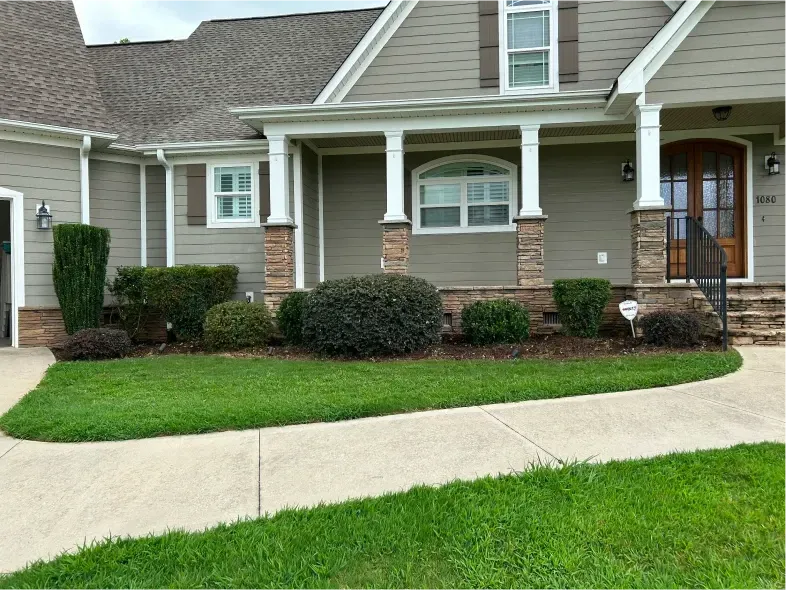 Tan house with green lawn, flower beds with bushes and a sidewalk.