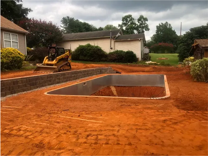 Construction site with a rectangular hole dug in red soil. A retaining wall and bulldozer are also visible.