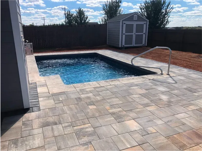 Small backyard pool with stone patio, shed, and blue sky.