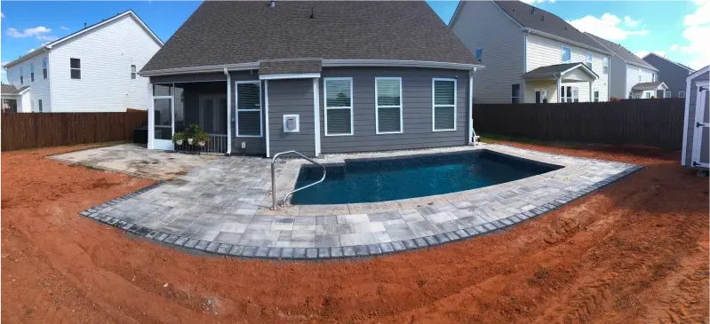 Backyard with a swimming pool, pavers, and a gray house. Red dirt surrounds the pool area.