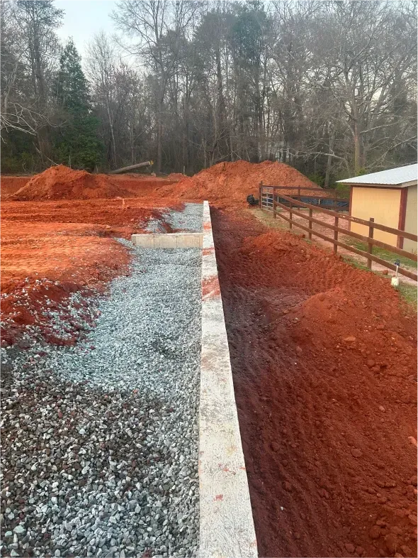 Red dirt and gravel separated by a concrete barrier, with large mounds of dirt in the background near a wooden fence and trees.