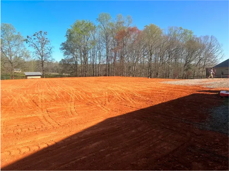 Cleared red dirt lot with tire tracks, trees in the background, bright blue sky.