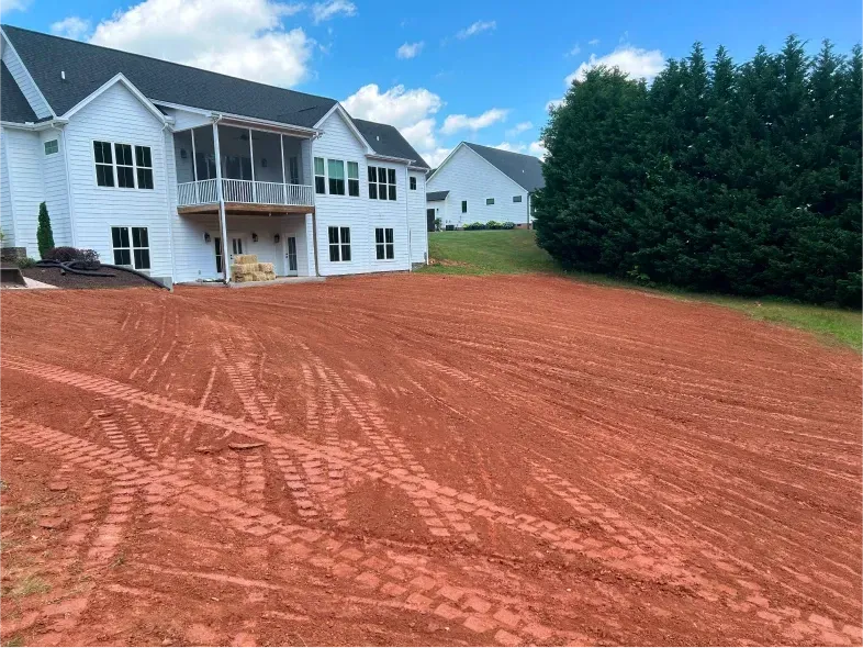 Red-dirt yard with tractor tracks, behind a white house and near a stand of green trees under a blue sky.