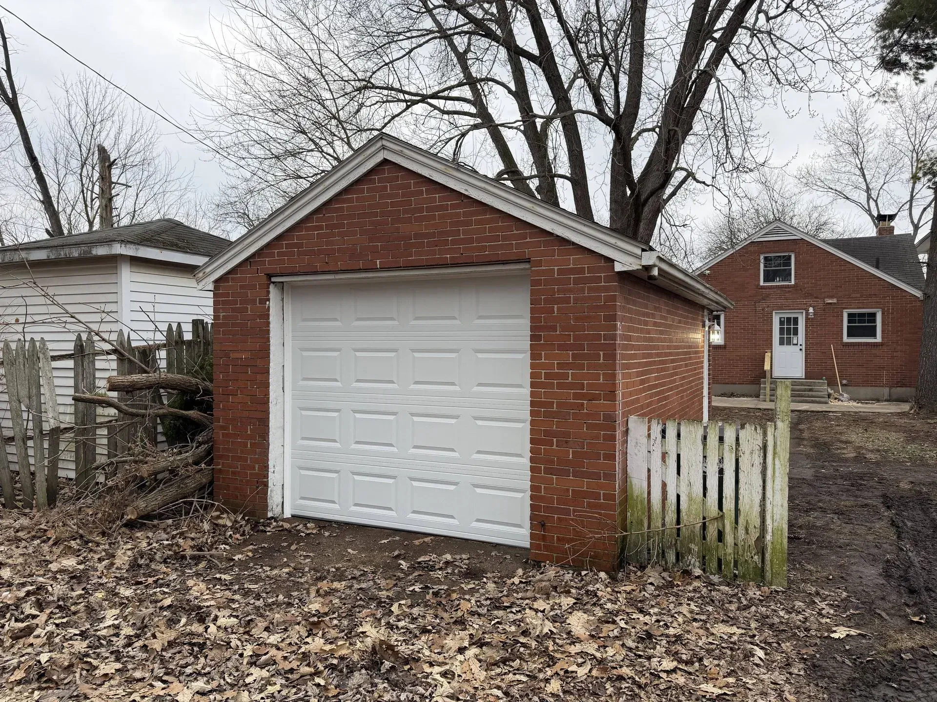 Small red brick garage with a white door beside a fenced yard, with leaf-covered ground and bare trees.