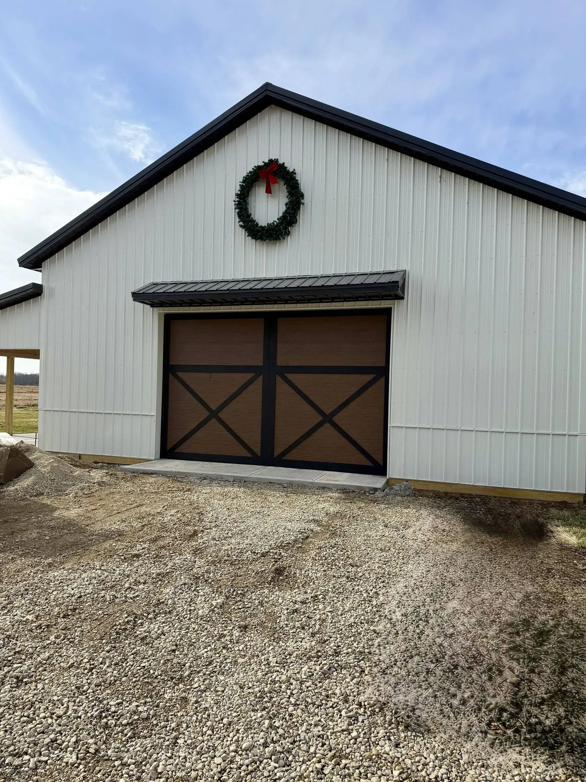 White barn with brown double doors and a Christmas wreath on the front, set on a gravel driveway.
