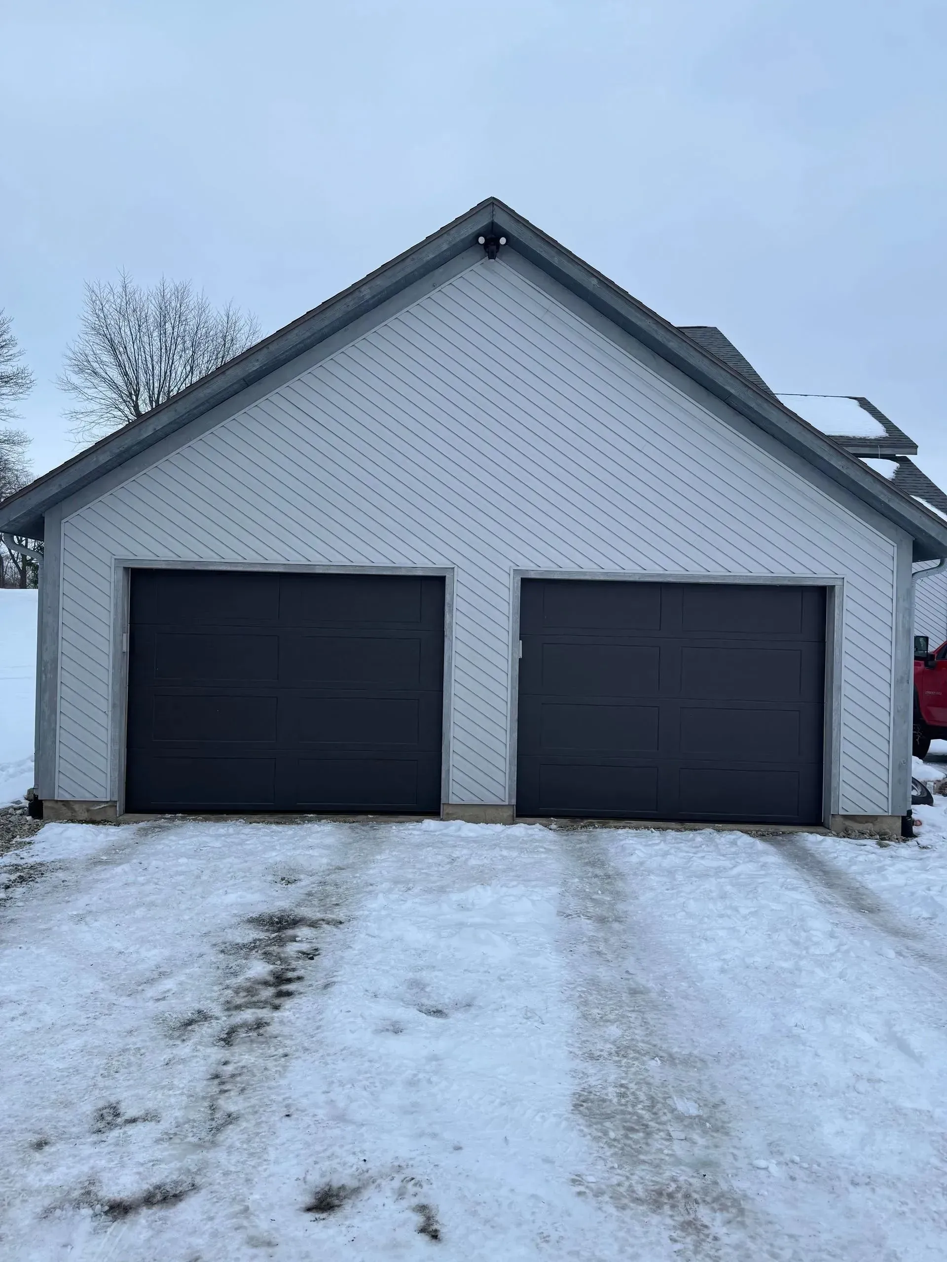 Snowy driveway leading to a white garage with two dark doors and a steep roof