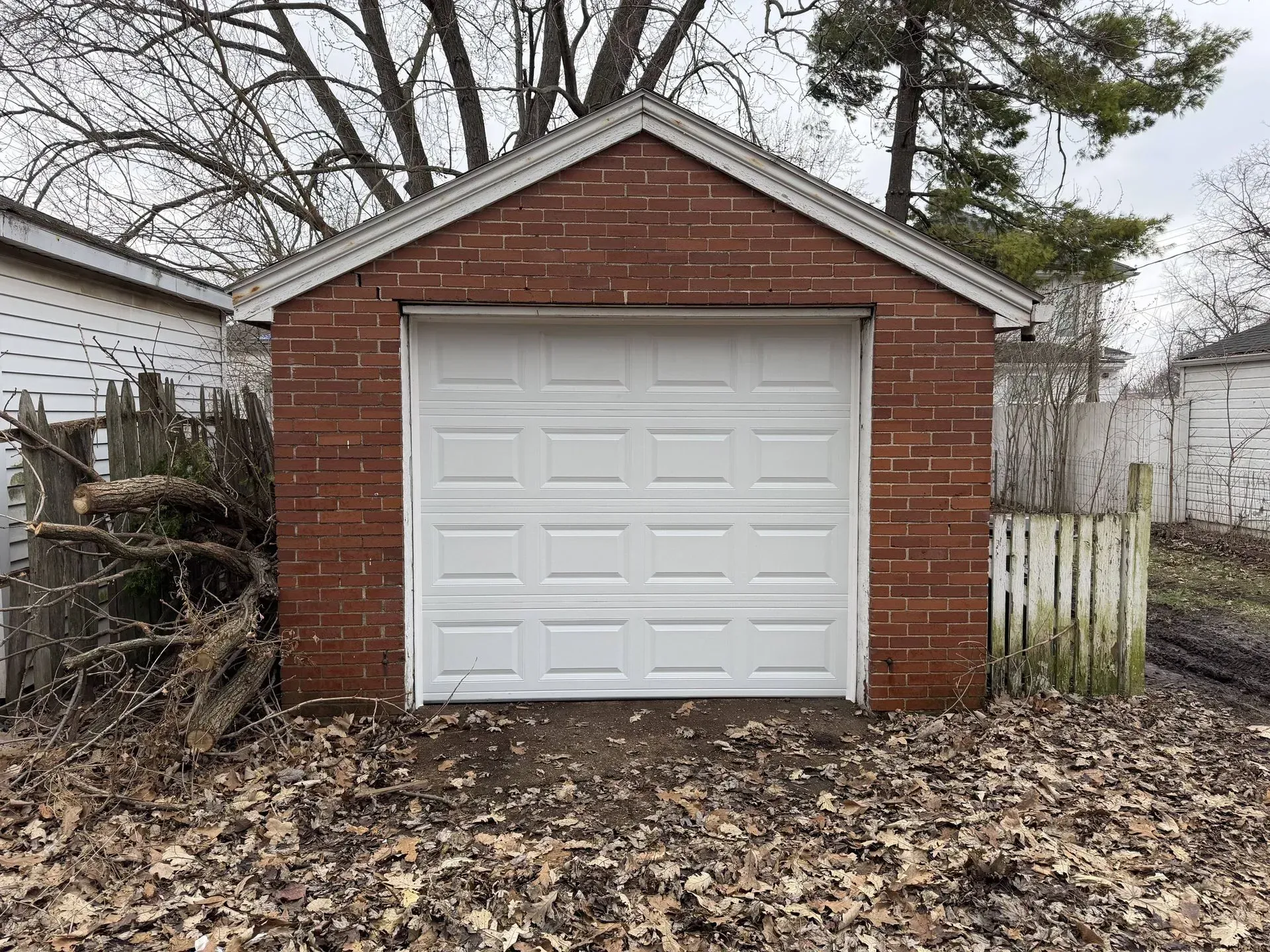 Small red brick garage with white door, surrounded by bare trees and fallen leaves in a backyard.