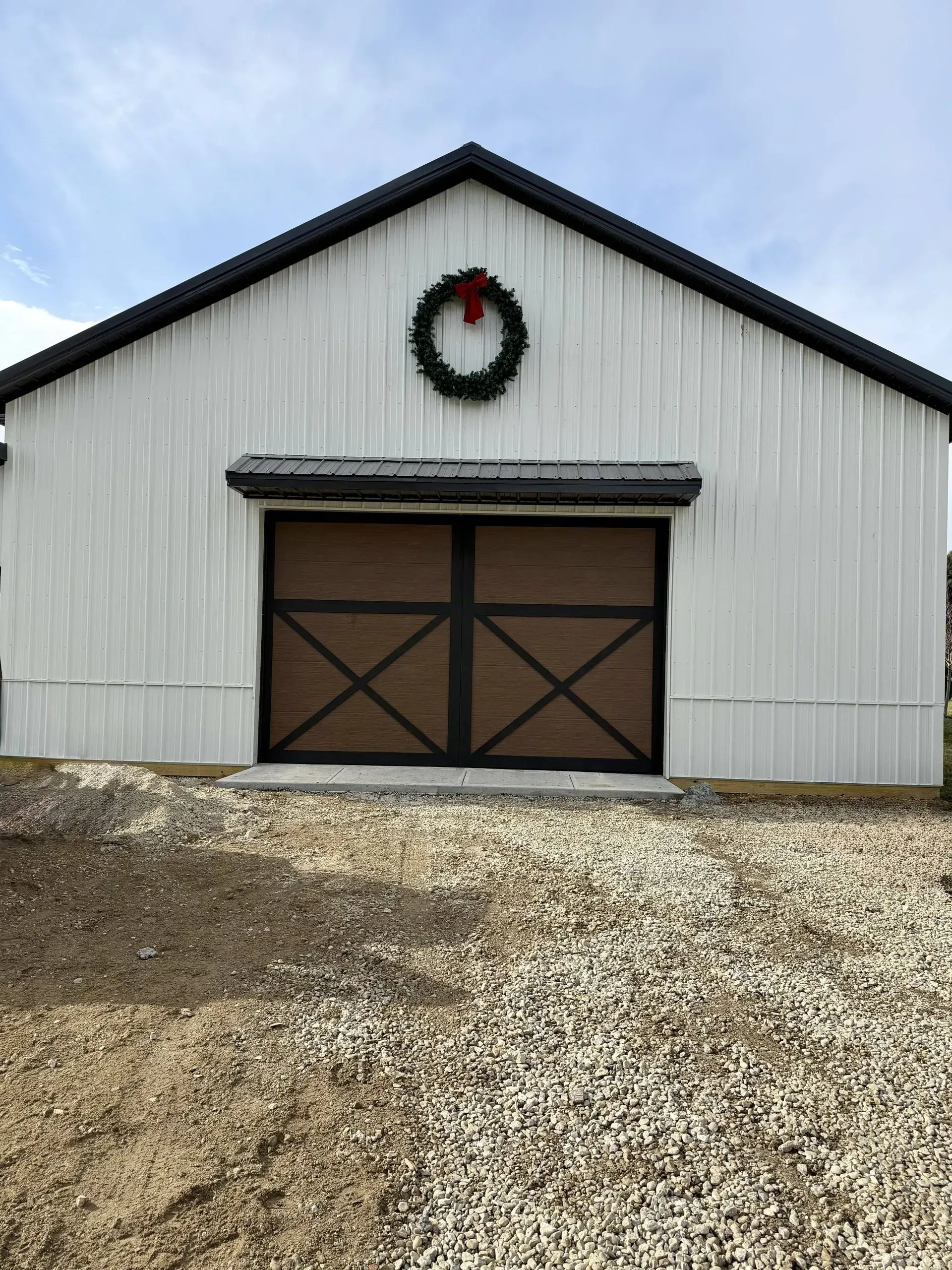 White barn with brown double doors, a wreath above, and a gravel driveway under a cloudy sky