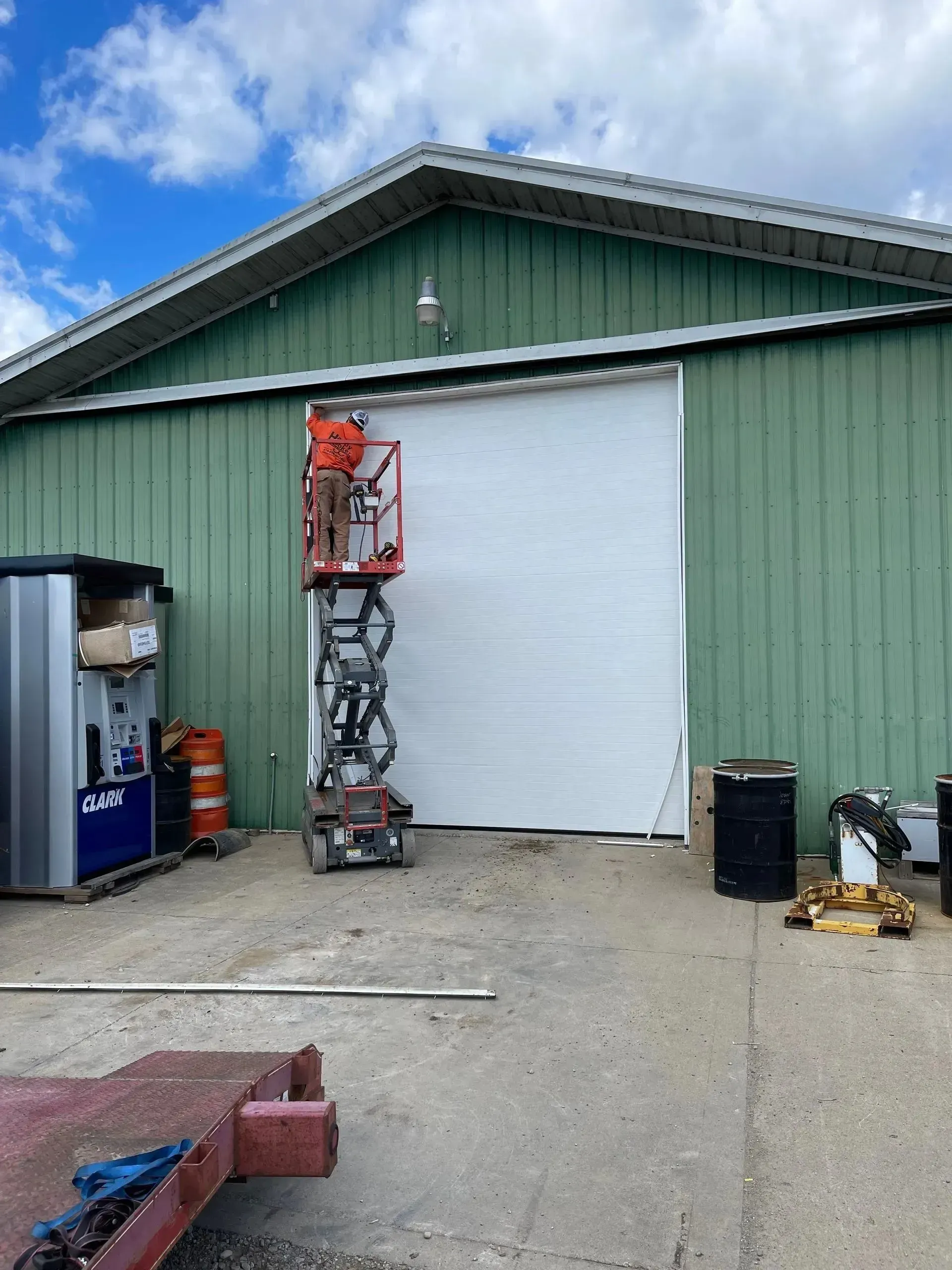 Green warehouse with a large white roll-up door, utility equipment, and a lift near the entrance under a blue sky