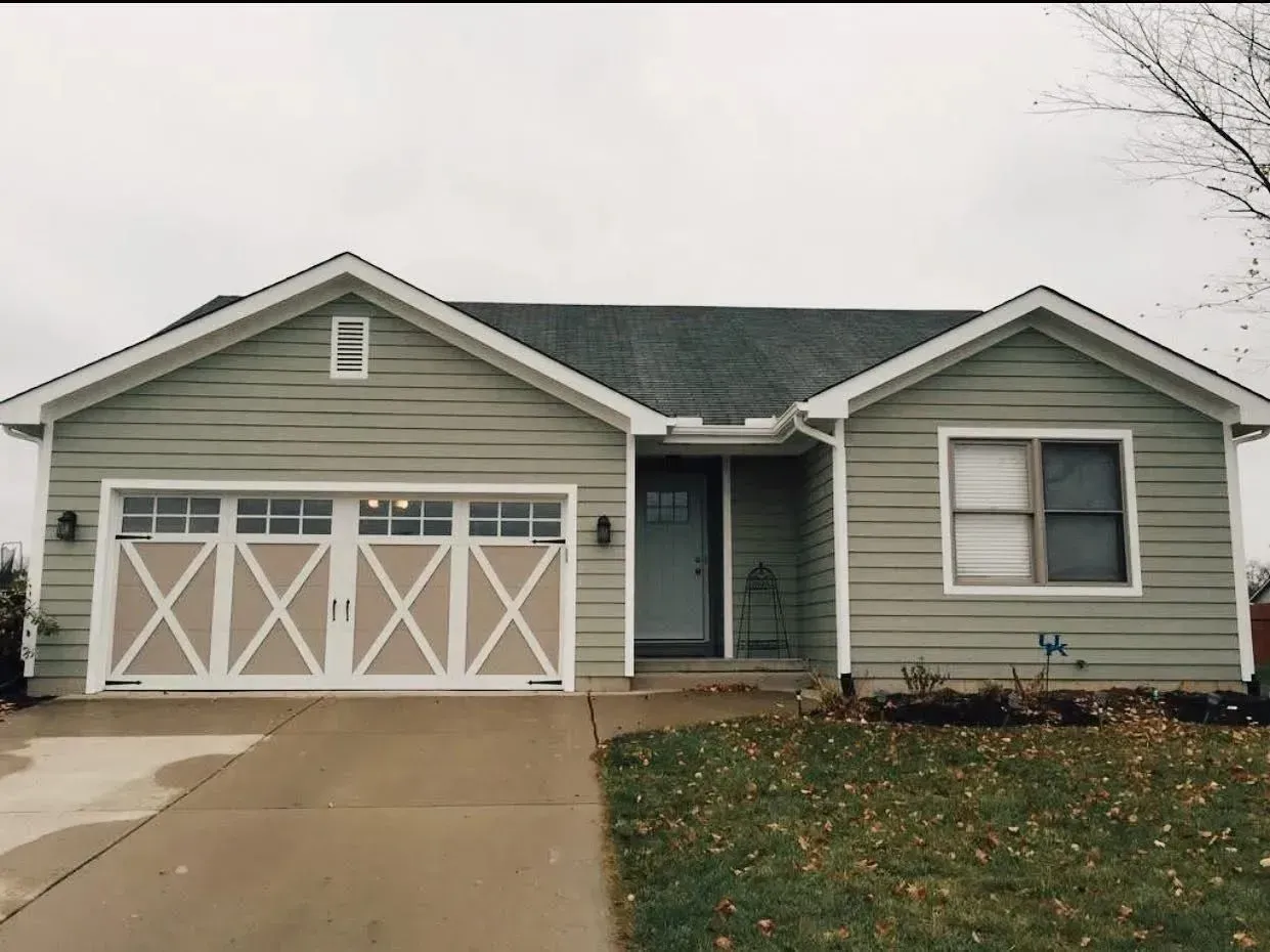 Single-story gray house with white garage doors, front porch, and a driveway on an overcast day