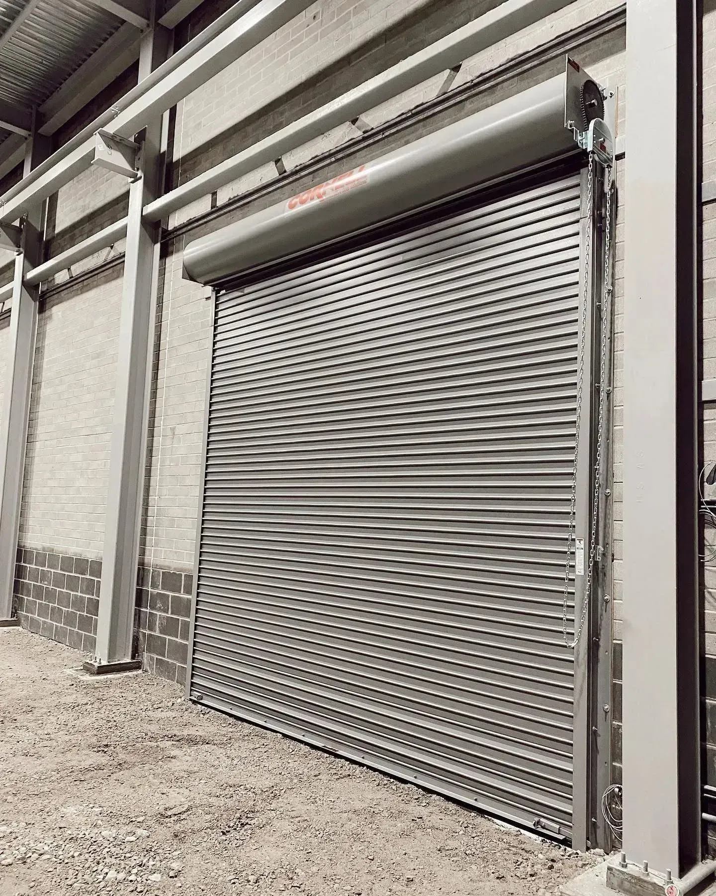 Gray metal roll-up warehouse door in an industrial building with gravel ground.