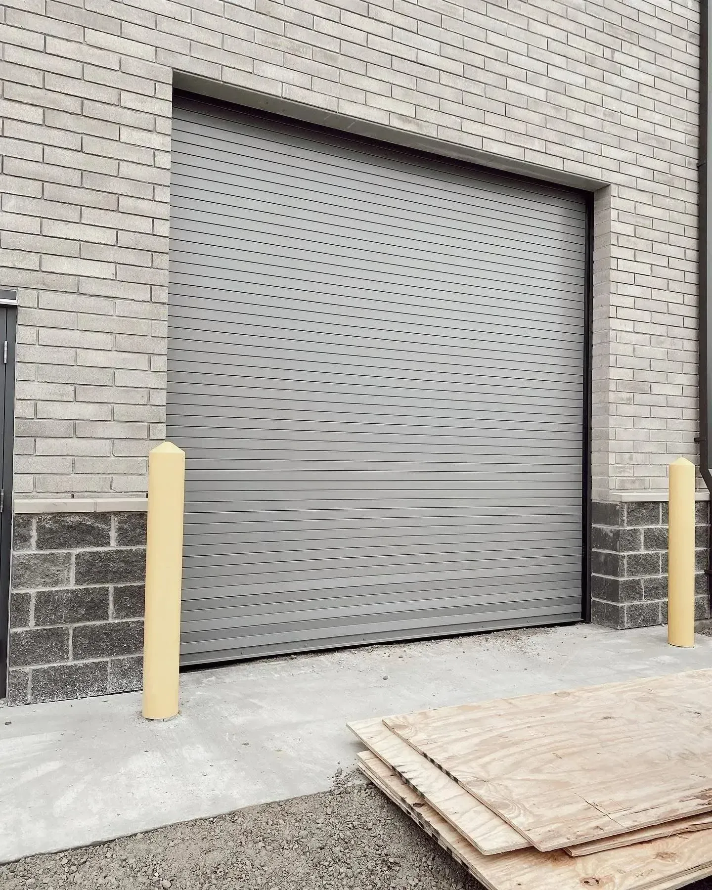 Gray roll-up loading dock door in a brick wall, flanked by yellow bollards.